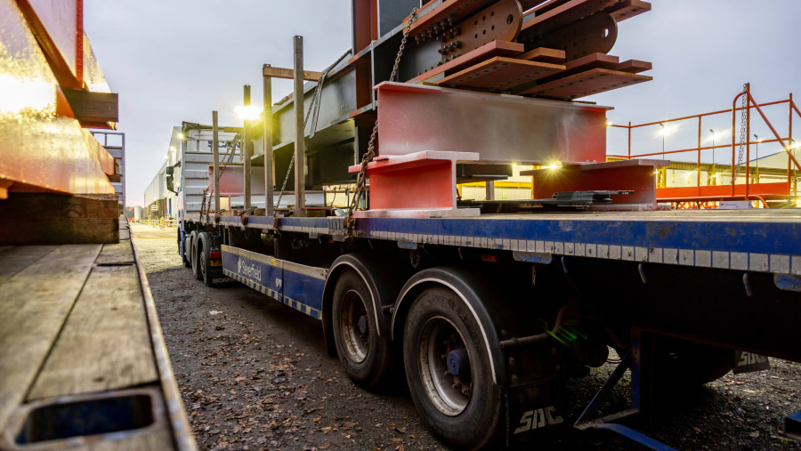 A long, blue flatbed semi-truck trailer loaded with large, rust-colored and gray steel beams. The truck is parked on a gravel and dirt lot, with a warehouse or industrial building visible in the background. The low angle of the photo emphasizes the size of the truck and its cargo.