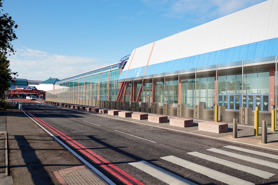 A temporary EnviroFence PRISON™ system with anti-climb mesh panels and concrete ballast blocks is set up in front of a modern airport terminal building. The fence runs parallel to a paved road with a pedestrian crosswalk in the foreground. The building features large windows and a blue and white facade, and the sky is clear and blue.