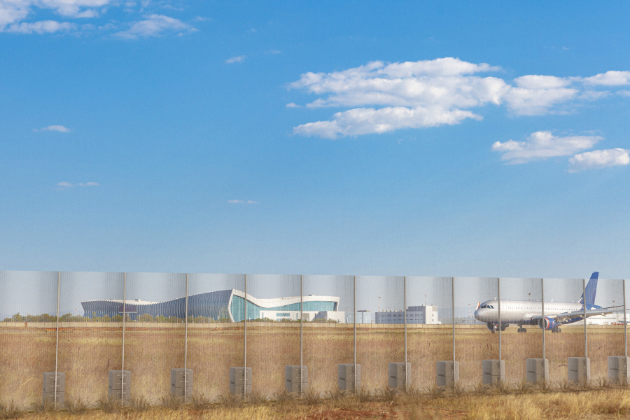 A low-angle view of a high-security temporary fence with an anti-climb mesh and concrete ballast blocks. The fence runs along the bottom of the frame, separating the viewer from a tarmac with a large commercial airplane visible on the right. In the background, a modern airport building with a curved, futuristic design is visible under a blue sky with scattered clouds.
