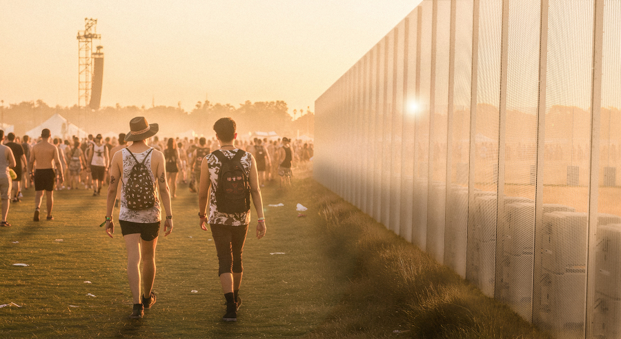 A long, high temporary fence made of metal mesh panels stands on the right side of a grassy field. The fence reflects the warm, bright light of a setting or rising sun. On the left, two people with backpacks walk away from the camera, and many other people are visible in the background, suggesting a festival or outdoor event setting. A tall speaker tower and several white tents are also visible in the distance