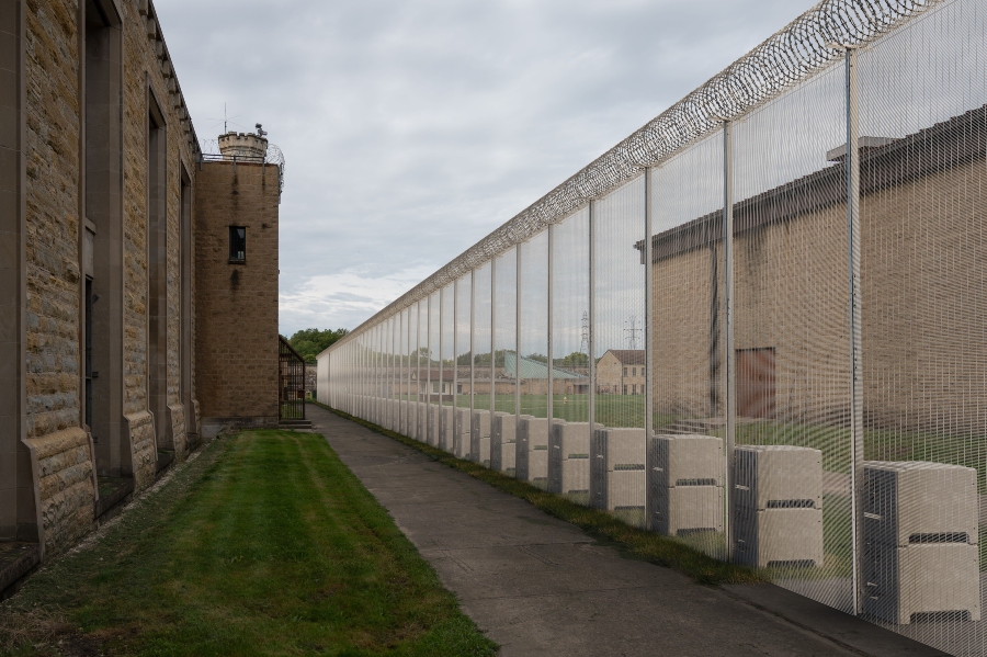 A tall, temporary security fence made of anti-climb mesh panels and supported by large, stacked concrete blocks lines a paved walkway. On the left is a large, light-brown building with a round turret visible in the distance. Another building is on the right, and the area is bordered by a grassy lawn and an overcast sky. There is razor wire on the top of the security fence.