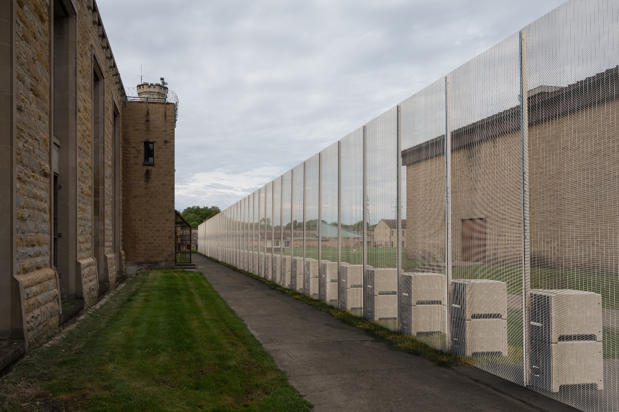 A 5.2m high, light-colored EnviroFence PRISON™ temporary security fence with anti-climb mesh panels extends along the side of a tall, light-brown brick building. The fence is supported by a series of gray ballast blocks and runs parallel to a paved path and a grassy area, with another building visible on the right. The sky is overcast.