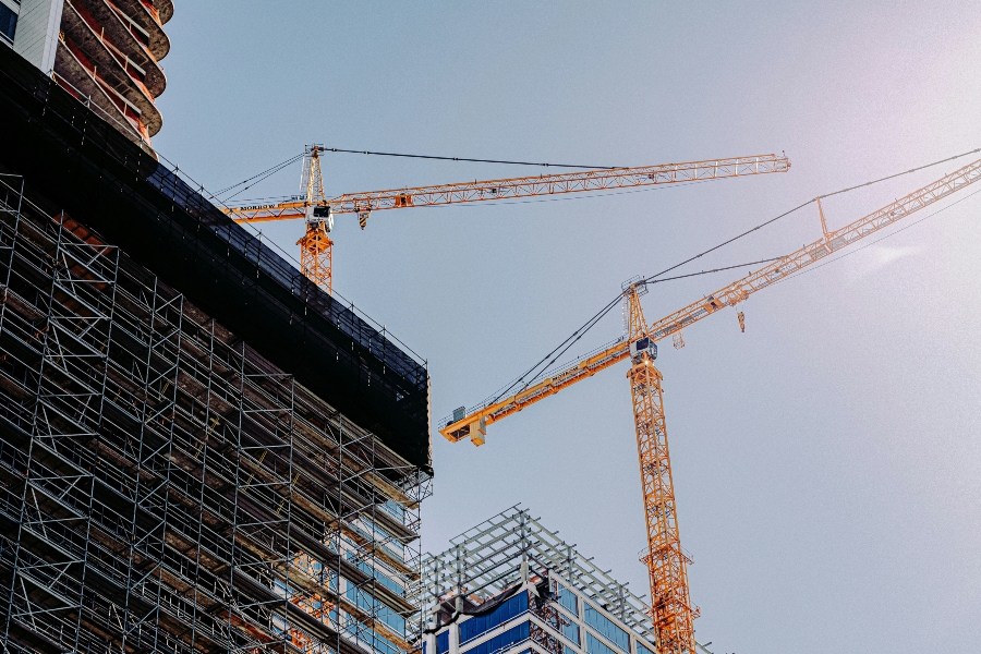A low-angle shot of a construction site showing the tops of two buildings and two large yellow cranes against a clear, pale blue sky. The building on the left is covered in scaffolding and dark netting, while the one on the right is a modern glass structure with a skeletal steel frame on top