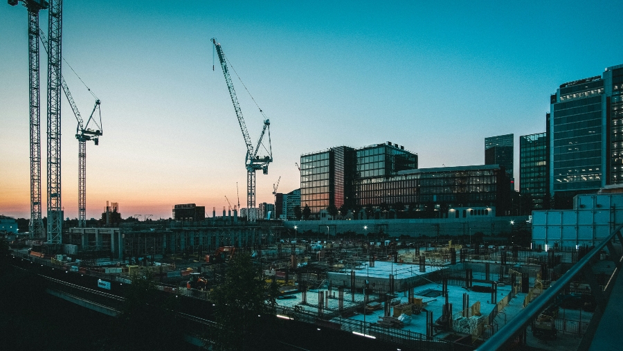 A wide-angle, low-light shot of a bustling construction site at dusk. In the foreground, the ground is torn up with exposed foundations, concrete forms, and construction materials. Several large cranes with extended booms dominate the left side and center of the frame, towering over the work in progress.