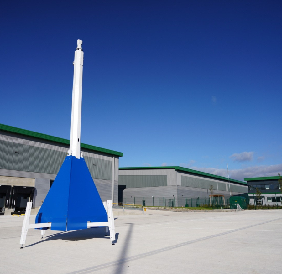 A blue and white mobile CCTV tower stands in an empty paved lot in front of a modern industrial building with a green roof, under a clear blue sky.
