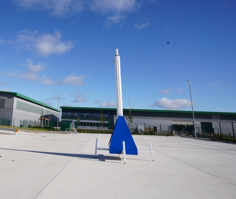 A close-up shot of the top of a white CCTV mast against a clear blue sky, showing a camera unit and other equipment.