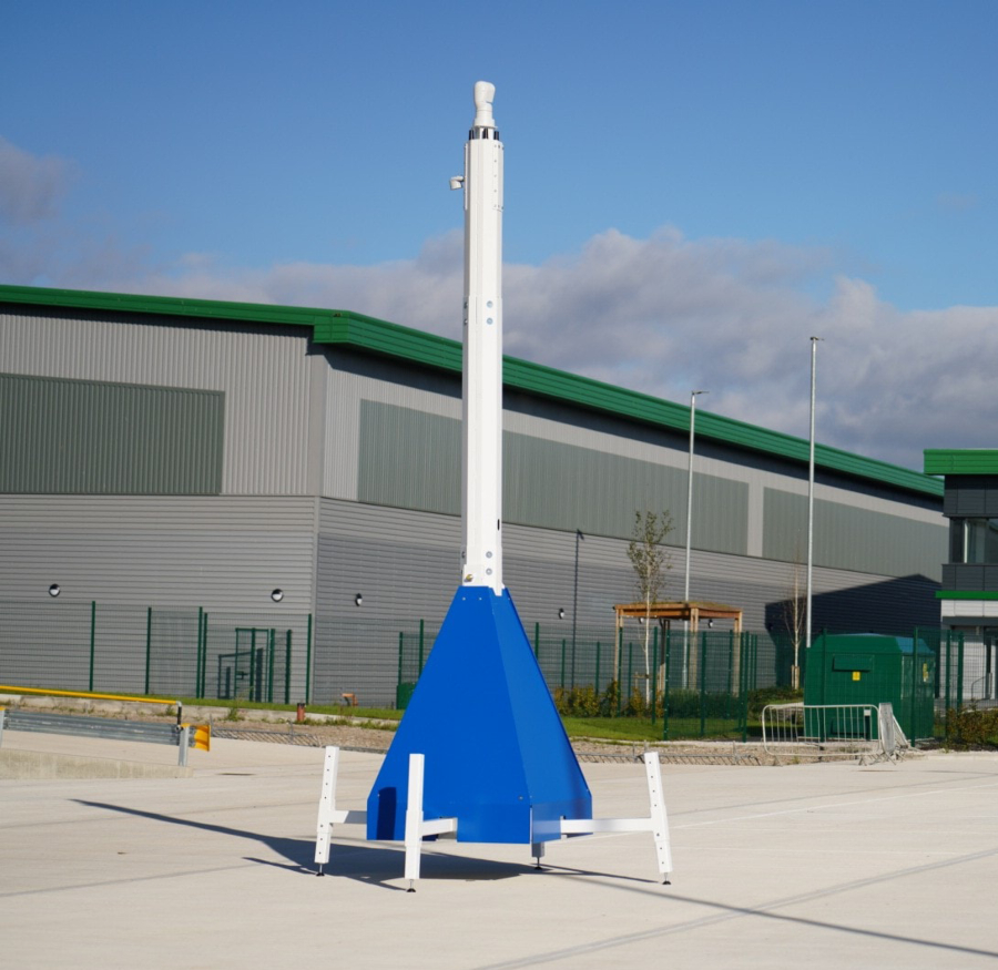 A front-on, eye-level shot of a blue and white mobile CCTV tower standing on a paved lot. The tower's camera mast is fully extended, and a large, modern industrial building is visible in the background under a blue sky.