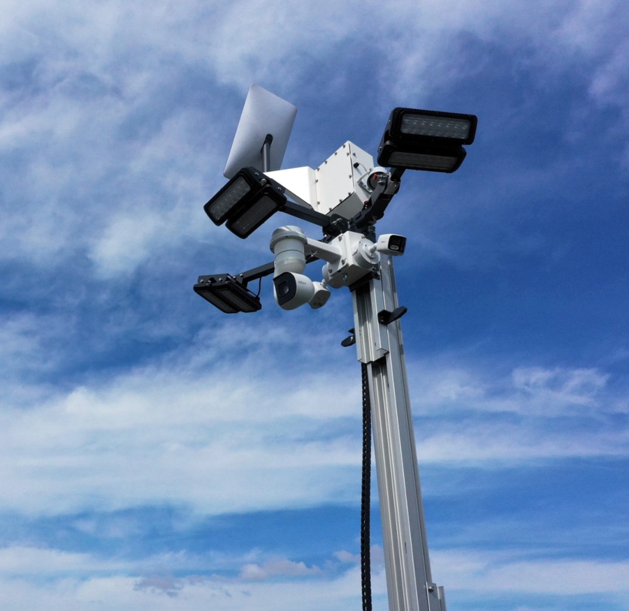 A low-angle shot of the top of a metallic CCTV mast, featuring multiple cameras and two black floodlights mounted on a white box, all set against a dramatic blue sky with scattered clouds.