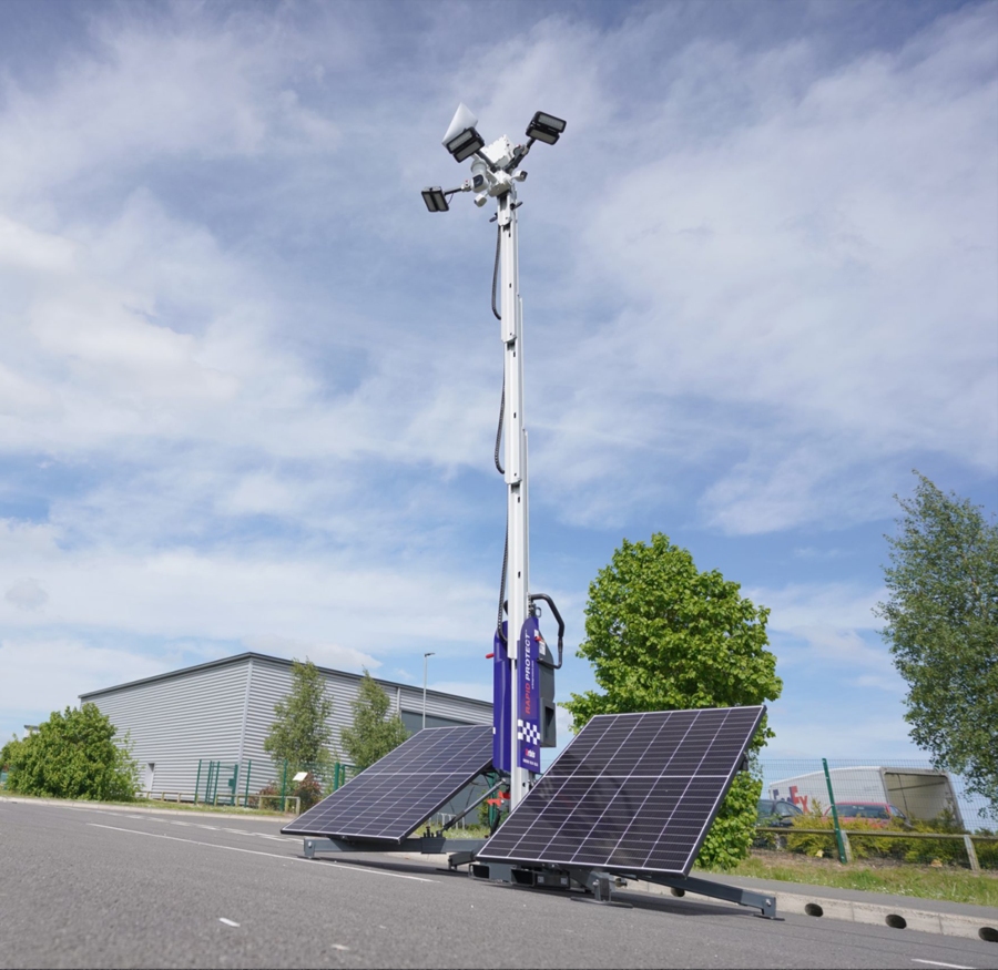 A tall, silver mobile CCTV tower with a blue base stands on a paved road, powered by two large solar panels on either side. In the background, there are industrial buildings, trees, and a partly cloudy blue sky.