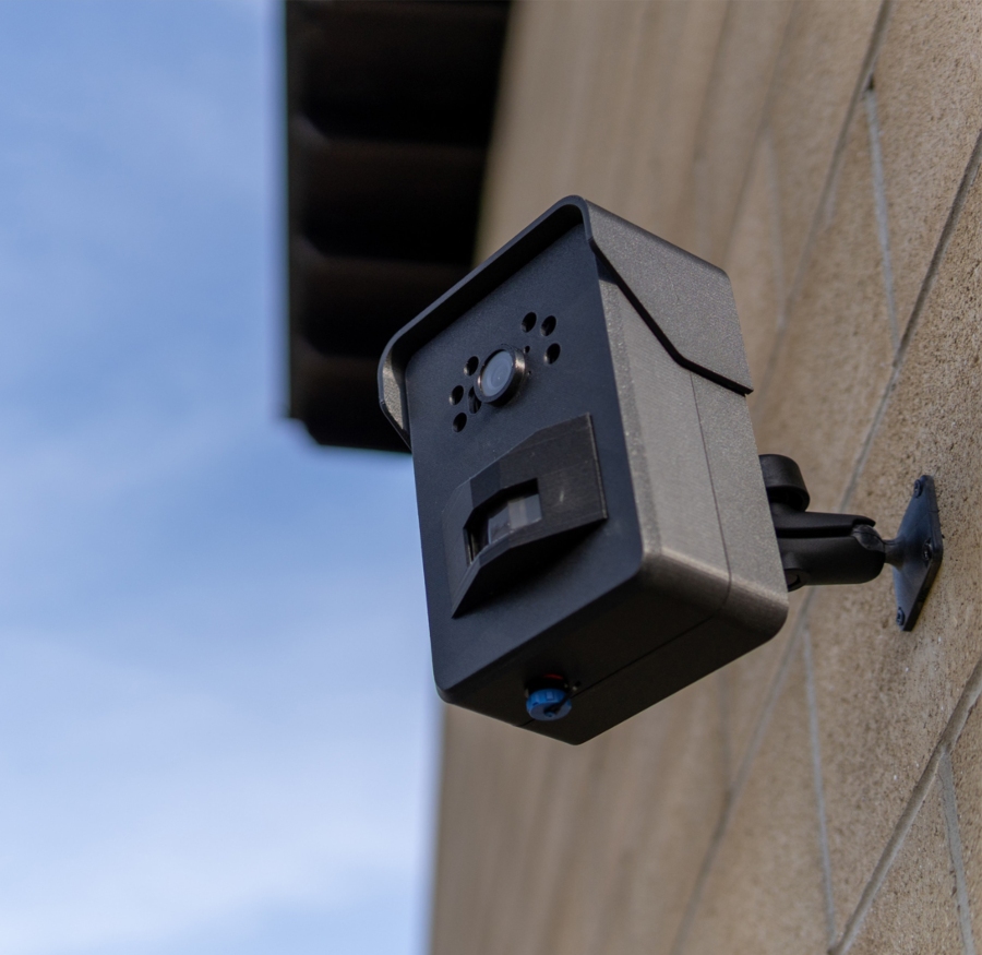 A close-up of a small, black CCTV camera mounted on a textured tan wall, with a clear blue sky in the background. The camera has a lens at the top, a rectangular sensor in the middle, and a blue light at the bottom.