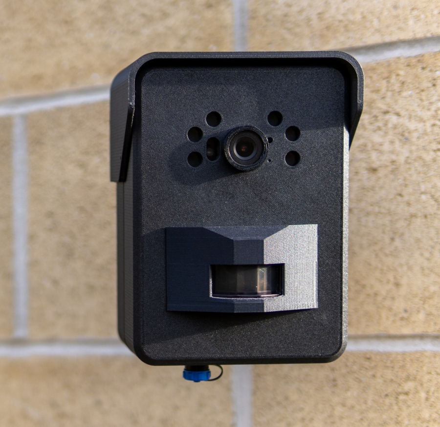 A close-up, head-on shot of a small, black CCTV camera mounted on a beige brick wall. The camera has a main lens at the top, a rectangular sensor below it, and a small blue element at the bottom.