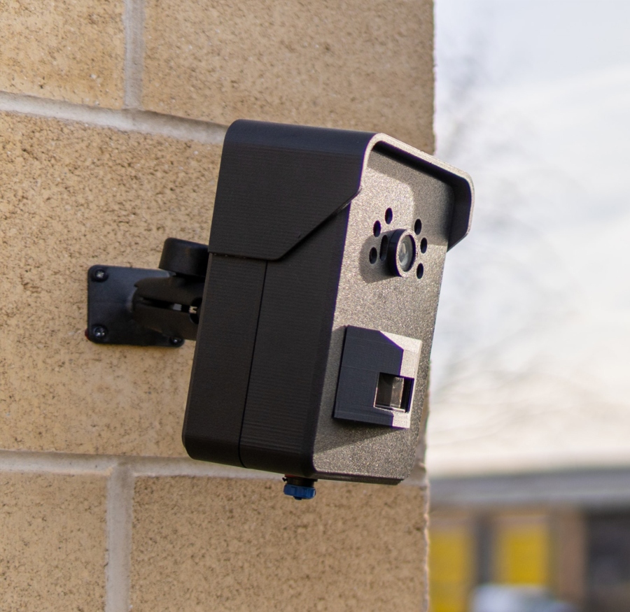 A side-view, close-up shot of a small, black CCTV camera mounted on a beige brick wall. The camera is angled slightly downwards, and a blurred background shows a building and a light sky