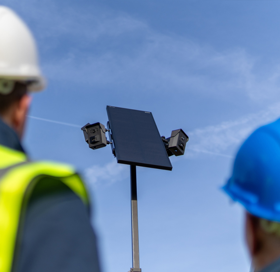 Two individuals wearing hard hats and safety vests are blurred in the foreground, looking up at a CCTV camera system mounted on a pole. The system includes a solar panel and two black cameras, set against a bright blue sky with wispy clouds.
