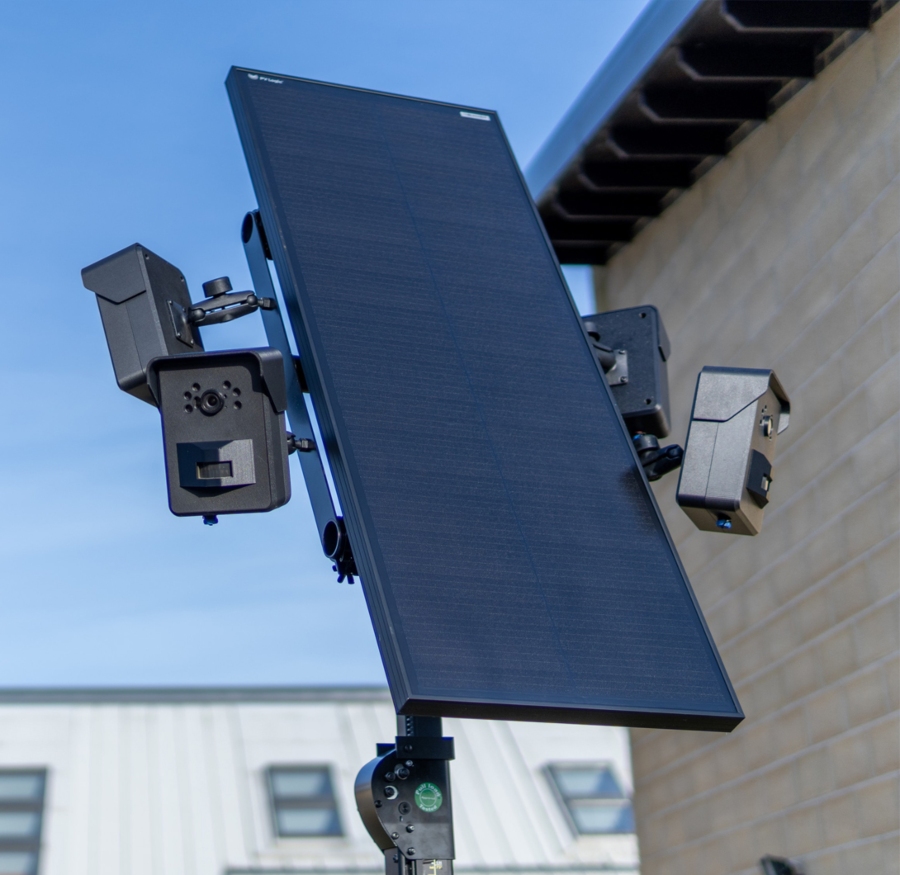 A close-up of a solar-powered CCTV system mounted on a pole, featuring a large black solar panel flanked by two smaller black camera units. The system is positioned outside a building with beige textured walls under a blue sky.