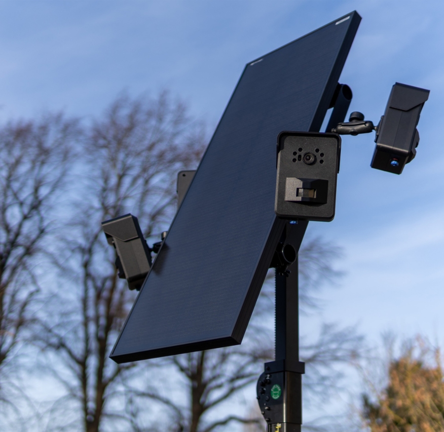 A mobile, solar-powered CCTV system stands outdoors, featuring a large black solar panel with two cameras mounted on either side. The backdrop is a clear blue sky and a row of bare trees