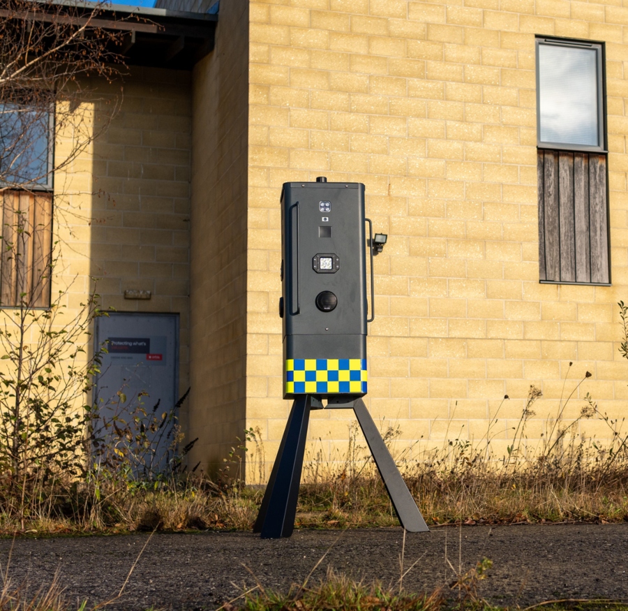 A dark gray mobile security camera unit, with a yellow and blue checkerboard pattern on its lower half, stands on a tripod in front of a tan brick building.