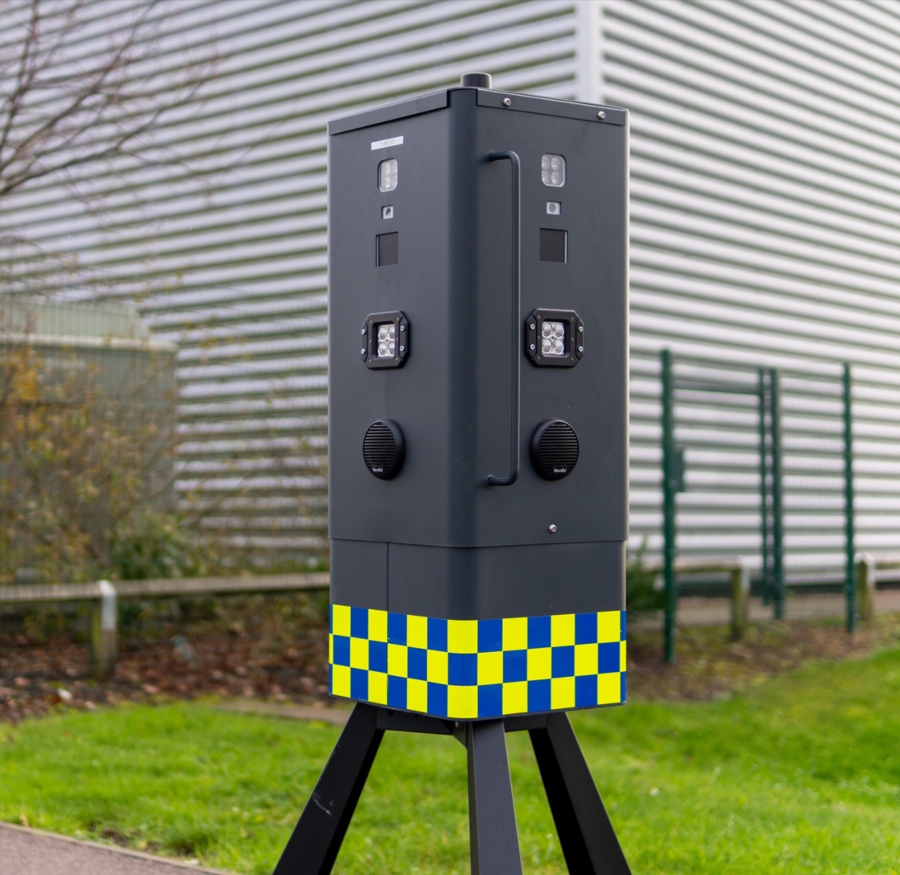 A large, black mobile CCTV unit with a yellow and blue checkerboard pattern on its lower half stands on a tripod. The unit has multiple lenses, lights, and speakers on its front panel. A modern building with gray corrugated metal siding is visible in the background