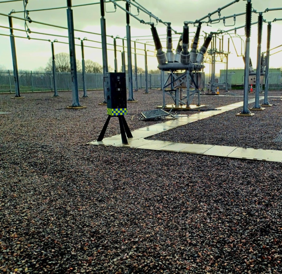 A mobile CCTV unit with a yellow and blue checkerboard pattern stands on a tripod in a gravel-covered electrical substation, surrounded by various power lines, transformers, and metal structures under a hazy, overcast sky.