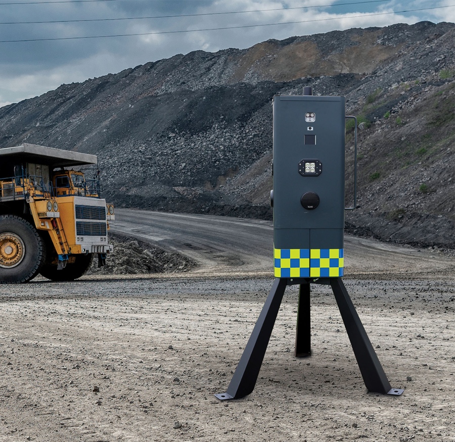 A portable, dark gray CCTV unit with a yellow and blue checkerboard pattern stands on a tripod in a dusty, open-pit mine or quarry. In the background, a massive yellow dump truck is partially visible next to a large pile of dark gray material.