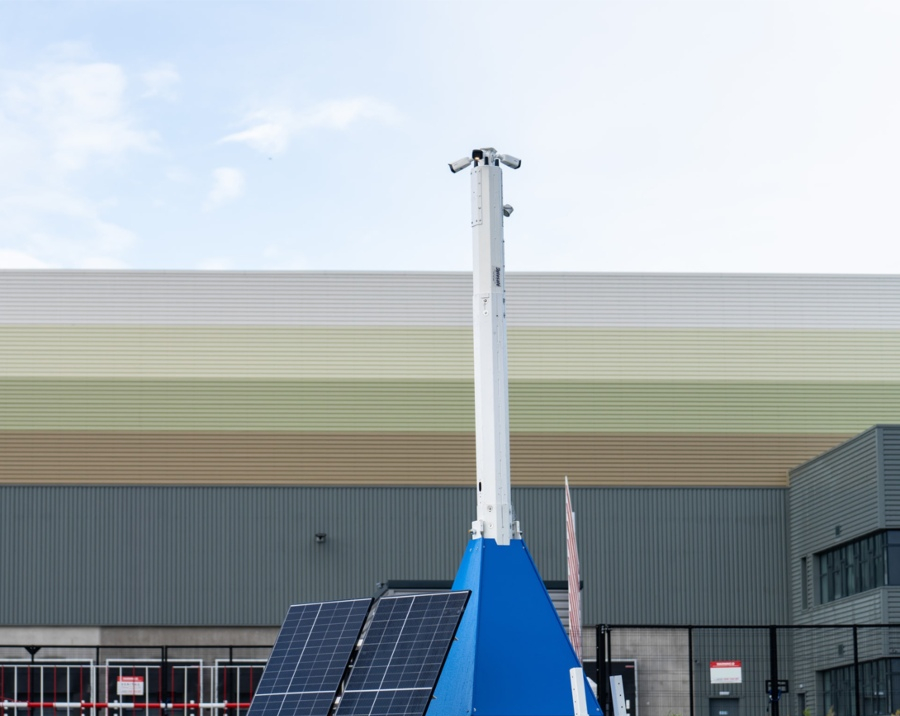 A front-on, eye-level shot of a blue and white mobile CCTV tower standing on a paved lot. The tower's camera mast is fully extended, and a large, modern industrial building is visible in the background under a blue sky.
