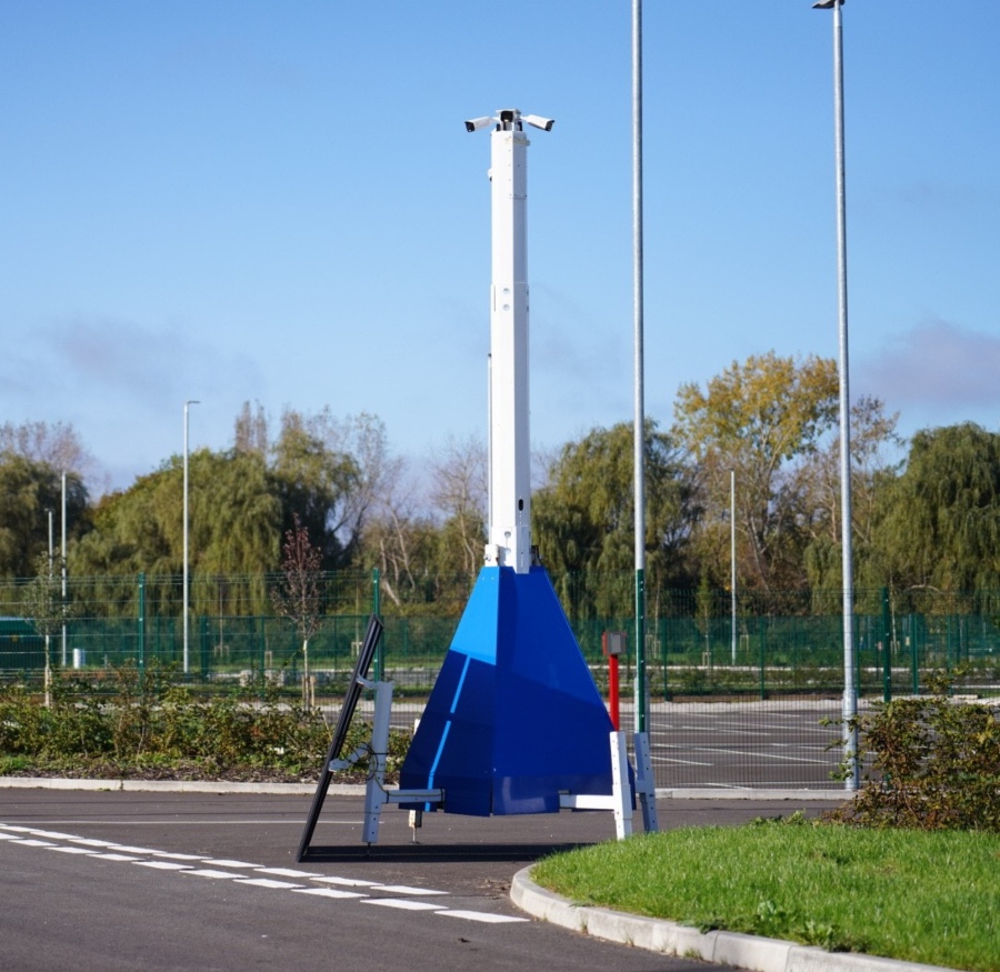 A large, blue and white mobile CCTV tower stands on a paved road, next to a grassy curb. The tower's mast is extended, and it is positioned in front of a chain-link fence and trees, under a blue sky with some clouds.