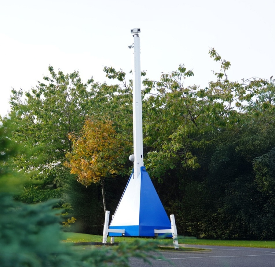 A tall, white mobile CCTV tower with a blue and white triangular base stands on a paved surface, surrounded by green trees and bushes under an overcast sky.